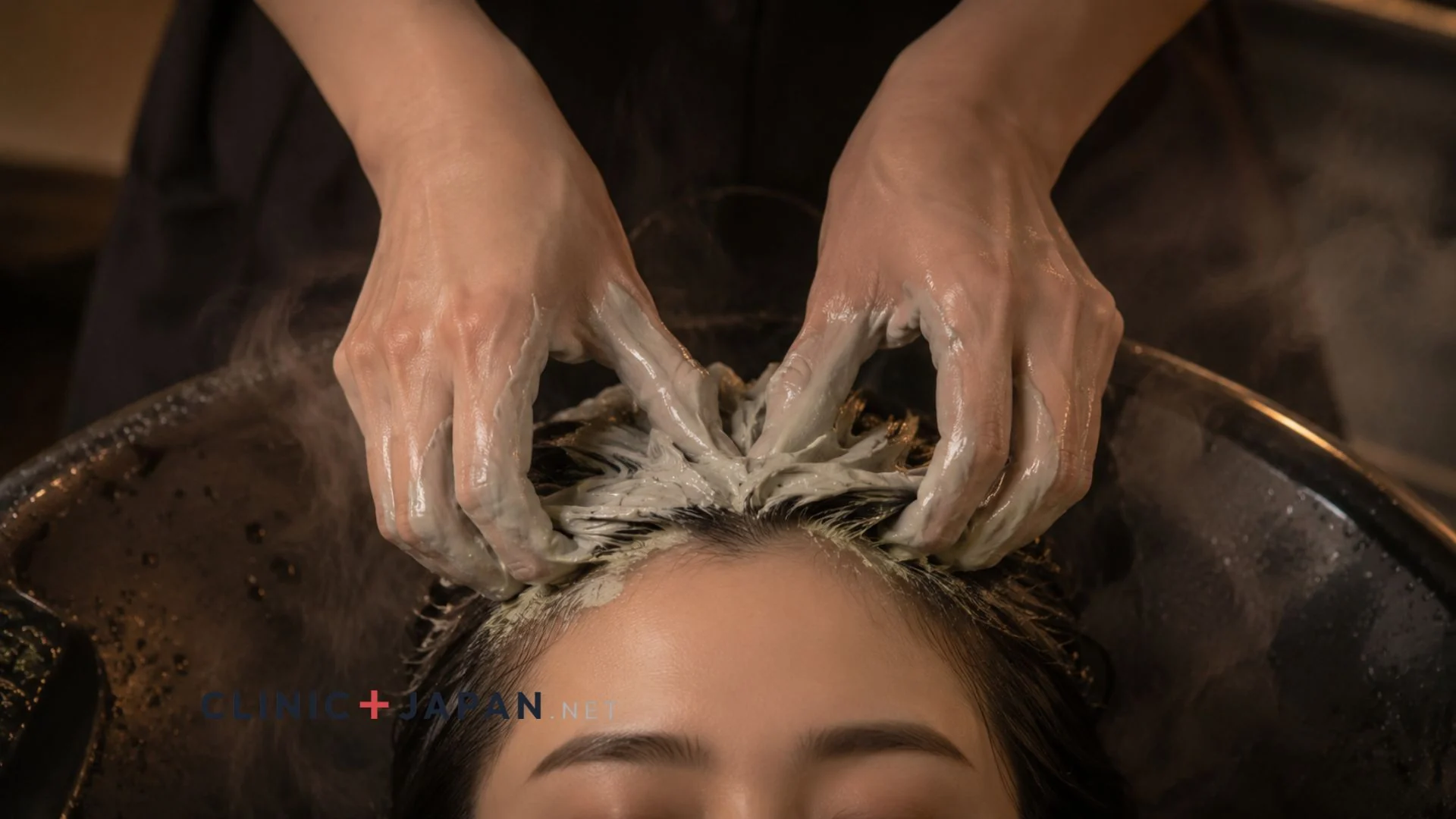 Therapist performing a wet Japanese head spa: gloved hands massaging clay-cream through the scalp of a client reclining in a shampoo basin