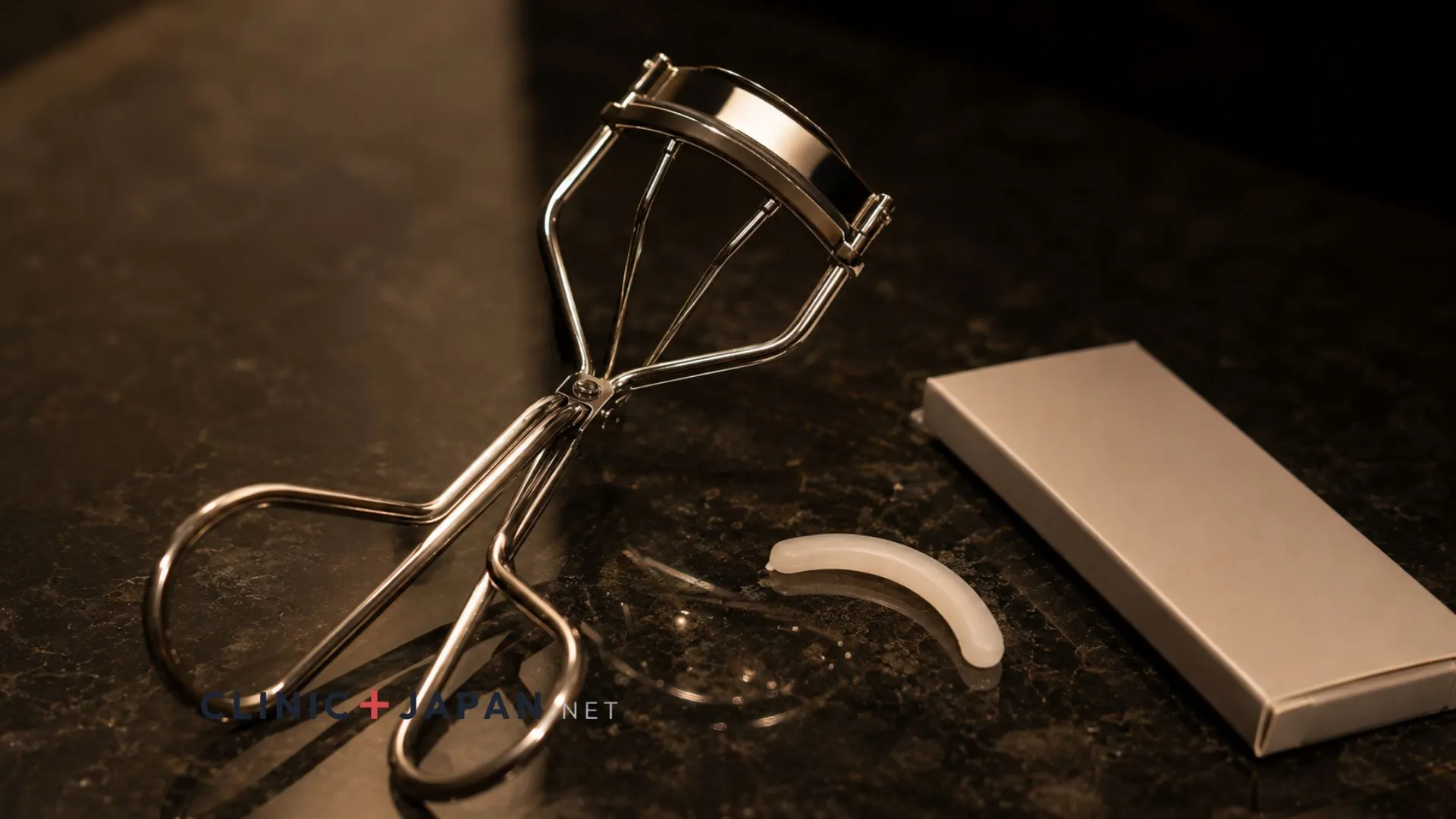 Metal eyelash curler resting on a dark department store counter beside a small silicone replacement pad and its box
